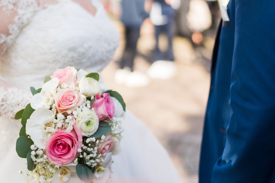 A close-up of a bridal bouquet featuring large pink roses, white ranunculus, and small white baby's breath, accented with green eucalyptus leaves. The bouquet is held by a bride dressed in a delicate 