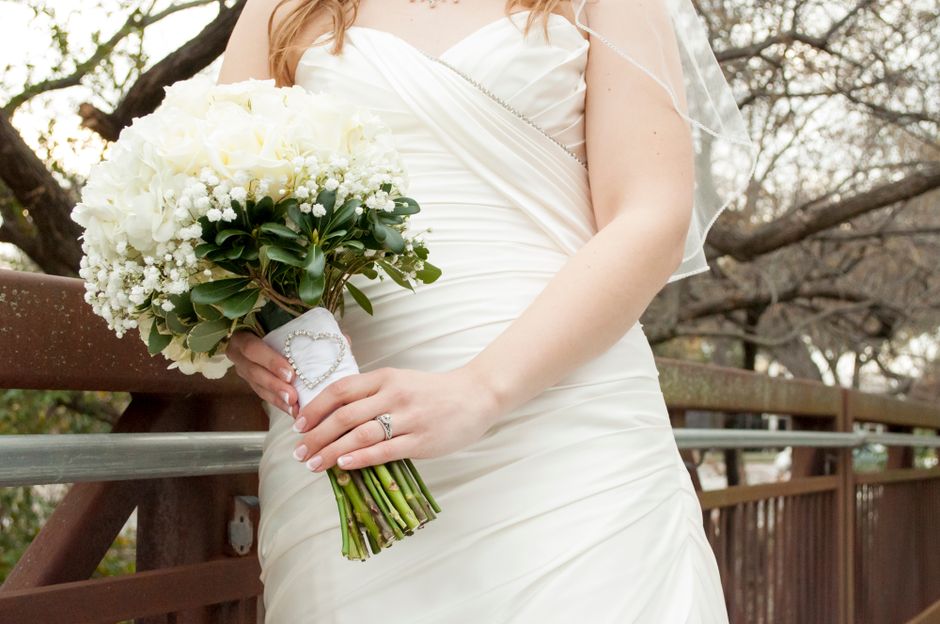 A bride in a white wedding gown holds a bouquet arrangement of fresh, predominantly white flowers, including roses and baby’s breath, with lush green foliage. The bouquet is wrapped with white silk 