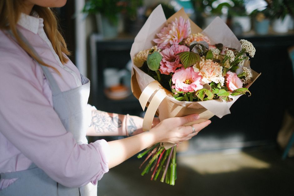 A person holding a bouquet of fresh flowers arranged with pink and cream blossoms, including carnations and chrysanthemums, wrapped in beige paper with a cream satin ribbon. The bouquet features lush 