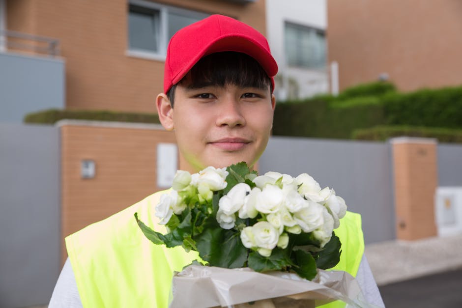 A young man with light skin, dark hair, and a gentle smile is holding a bouquet of white flowers, including roses and greenery, wrapped in white paper. He is wearing a red cap and a bright yellow high