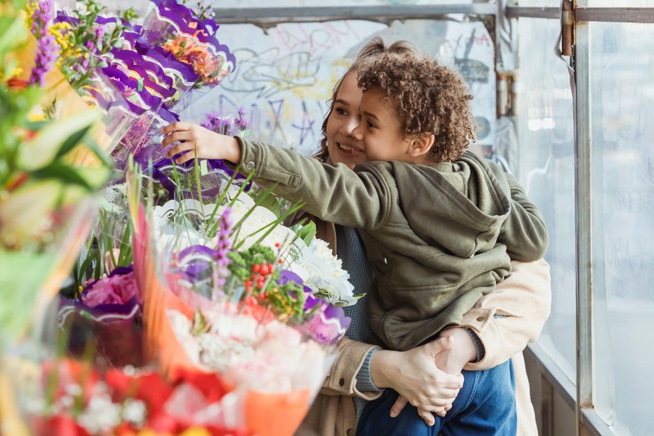 A young boy with curly hair and a cheerful expression is held in the arms of an adult, both smiling, while selecting vibrant floral arrangements at a flower market or shop. The bouquet features a mix 