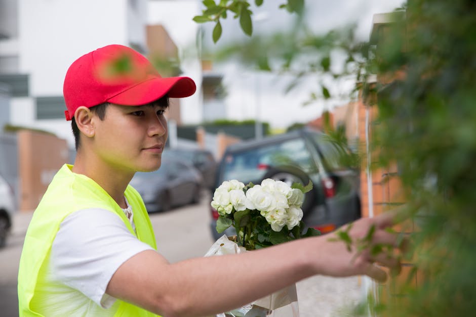 A young delivery person wearing a red cap and a high-visibility yellow vest is seen placing a floral arrangement at a residential doorstep during daylight. The bouquet features white roses and other w