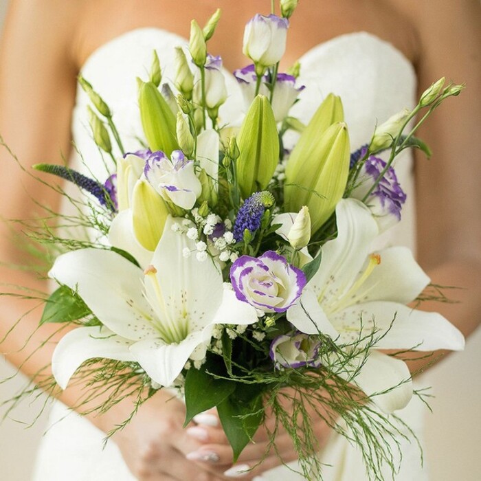 Bathed in serene morning light, this handcrafted bridal bouquet makes for an unforgettable moment in Hook. White Oriental lilies, some in full exuberant blossom and others in the quiet promise of unopened buds, dominate the arrangement, their silken petals marked with subtle speckles and prominent golden stamens. Lavish lisianthus blossoms, edged delicately in lavender purple, weave among them, mixing states of bloom and bud for dynamic texture. Deep purple spikes of Veronica lend vibrant colour and drama, while traditional white baby's breath floats amidst the larger florals, imparting an airy softness. Rich green foliage-big glossy leaves, unopened lily buds, and feathery fern-cascades from the base, framing the bouquet with a sense of movement and natural grace that recalls the local gardens and woodlands of Hook. The bride's manicured hands, painted in a soft, light shade, gently support the bouquet, their presence adding intimate humanity to the composition. Against the blurred backdrop of the bride's pristine white, strapless gown and bare shoulder, the flowers are spotlighted as a symbol of love and anticipation-romantic, fresh, and sophisticated. Available for bespoke wedding arrangements in Hook, this bouquet is a testament to local floral design and the start of a joyful journey.