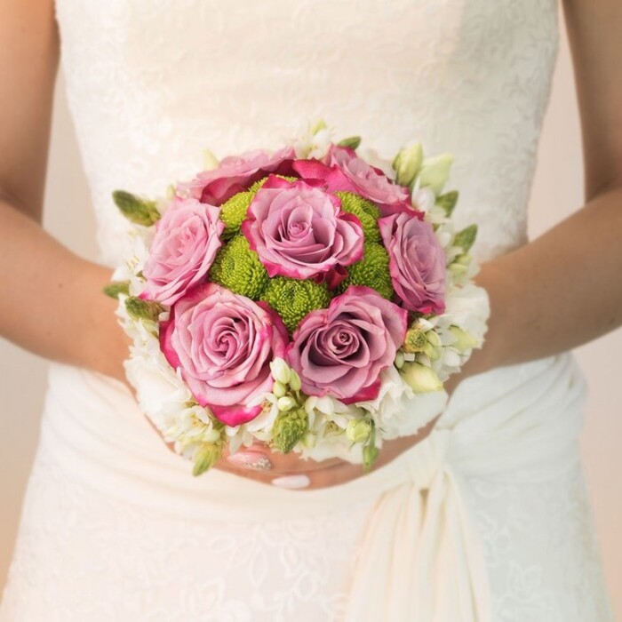 A loving glimpse into a Hook bridal morning reveals a compact, glowing bouquet, artfully held against an ivory gown adorned with exquisite lace-a familiar sight for those gathered at St. John's or strolling Hook's tranquil lanes. The focus is on a swirl of fully bloomed roses, each petal a painterly transition from faded mauve at the center to lively fuchsia edging, the effect both dynamic and tender. Between these standout roses, a cluster of fresh, lime-green button chrysanthemums forms a cheerful textured core, evoking memories of leafy afternoons in Hook's parks. Encircling the heart of the bouquet, petite clusters of white flowers-perhaps dainty freesia or spray roses, interlaced with slim green buds-bring purity and freshness, their delicate fragrance barely hinted in the morning air. The bride's forearms, lightly tanned and graceful, support the flowers with a gentle touch, nails polished in pale blush, catching the light that filters softly through a nearby window. Behind, the subtle detailing of the gown-lacework over the bodice and an understated drape at the hip-reflects a style both refined and welcoming, much like the spirit of Hook itself. This image is a celebration of artistry, capturing the harmonious interplay of colour, texture, and emotion, and serves as an inspiring example for couples seeking local, bespoke wedding florals that carry the joy and warmth of their community.