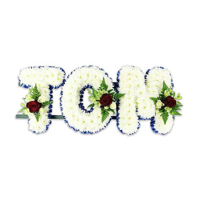 Bold yet tender, this personalised floral tribute spells 'TOM' in large, three-dimensional letters, the surfaces densely sheathed in delicate white chrysanthemum blooms that create a soft, tactile field of petals. The floristry technique is evident in the even, compact placement of each blossom, producing rounded forms that catch soft interior lighting and convey a sense of calm. Each letter is neatly edged with a vivid royal blue ribbon, the colour providing a refined border that crisply defines the shape against the white backdrop. Within each letter a formal spray is thoughtfully arranged: a single, rich red rose sits as the focal point in each cluster, its velvety layers suggesting deep affection, while smaller ivory spray roses and fine fern fronds surround it, adding sculptural contrast and a quiet green framework. A slim dark metal bar visible at the base stabilises the composition for upright display on easels or church stands. The overall aesthetic reads as a respectful, handcrafted memorial piece - visually clear, emotionally measured - and the faint fragrance of fresh roses and green leaf lends a discreet sensory presence appropriate for a funeral space. This style of bespoke name tribute is often chosen by families in Hook for services at the village church, local chapels, or delivery to funeral directors along Station Road and nearby Basingstoke Road, where it becomes a tangible gesture of sympathy and remembrance.