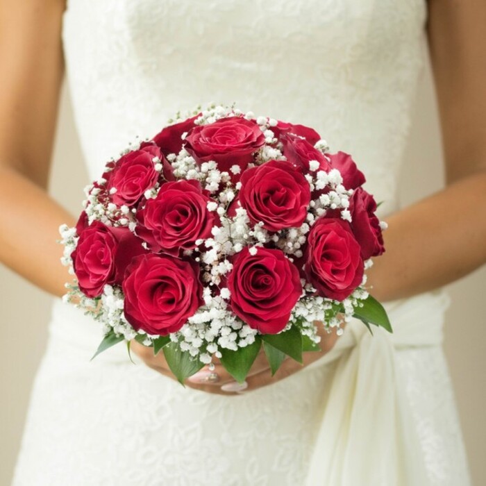 This evocative close-up centers on the hands of a bride from Hook, gently clasping a round and classic bridal bouquet rich with meaning and artistry. The bouquet radiates a deep, velvety red through its many roses, each bloom at a different stage of unfurling-some tightly coiled with anticipation, others luxuriantly open-embodying both the freshness and timelessness of true love. Between the roses, intricate clusters of pure white gypsophila-baby's breath-scatter like tiny clouds, softening the arrangement with their delicate spray. Lush green leaves, just visible at the base, suggest the bouquet has only recently been gathered, perhaps fresh from a morning delivery run along Sheephouse Road. The bride's artfully manicured fingers form an elegant support at the bouquet's base, their gentle hold suggesting both excitement and serenity. Her gown, visible in ivory-toned detail, features a pattern of lace or embroidery across the bodice, with a satin drape flowing gracefully to the side. All of this is set against a muted, creamy background that quietly fades from view, ensuring that every highlight-from the petals' velvet sheen to the bouquet's garden-fresh fragrance-draws the viewer in. Brides near Hook cherish this style for its understated romance and classic beauty, a testament to enduring love and local floral craftsmanship, perfect for a wedding day to be remembered.