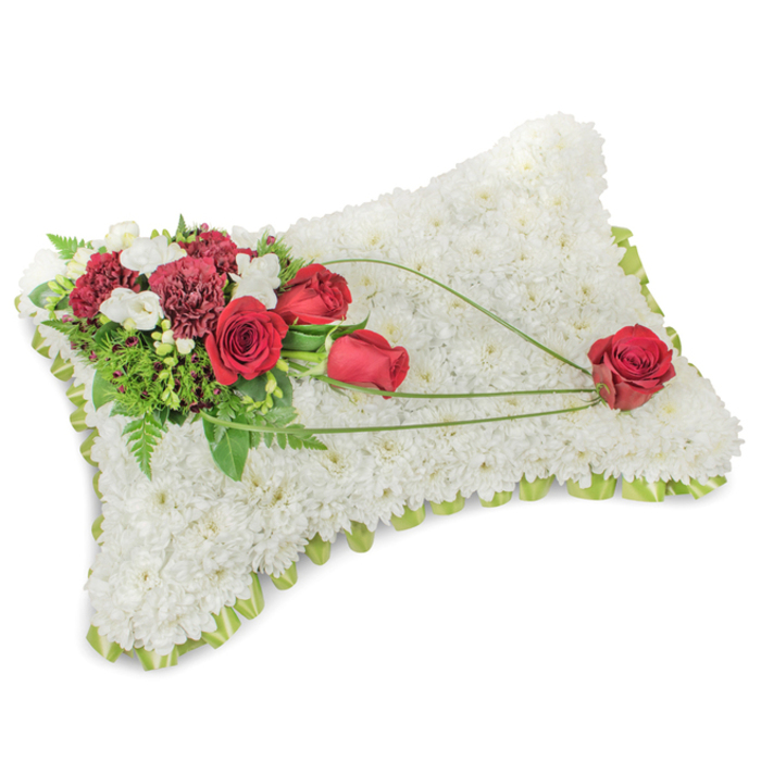 A tranquil, thoughtfully composed memorial cushion occupies the frame: the rectangular shape is meticulously formed from hundreds of small, immaculate white chrysanthemum blooms, packed tightly so the surface reads like a plush, snow-white textile. Along the slightly raised perimeter a faint thread of pale green-either satin ribbon or tender foliage-emerges between the blooms, softening the edge with a cool, fresh tone. On the upper-left quadrant a vibrant cluster of crimson and burgundy flowers creates an expressive accent: deep red roses in varied stages of bloom nestle against ruffled, dark-hued carnations and are brightened by petite white accent flowers, such as freesias or spray chrysanthemums, which scatter points of gentle light across the composition. Lush green foliage, from broad leaves to fine fern fronds, forms a verdant backdrop that heightens the reds' richness. From the main spray, three elegant lines of bright green bear-grass or similar slender foliage sweep diagonally across the chrysanthemum field toward the lower-right corner, establishing a flowing rhythm. At that corner a single, perfectly shaped red rose appears to rest, cradled by the curving stems-a solitary symbol of love and memory. The textures are tactile in description: velvet petals, feathery fern, and the soft matte of chrysanthemum heads; the scent, subtle and clean, blends gentle rose with fresh green notes. This classic sympathy arrangement reads as a calm, respectful tribute-suitable for a funeral service, chapel presentation, or a private commemoration at home. Prepared with local sensitivity in Hook and mindful of nearby communities around the M3 corridor, it expresses comfort, honour, and enduring affection.