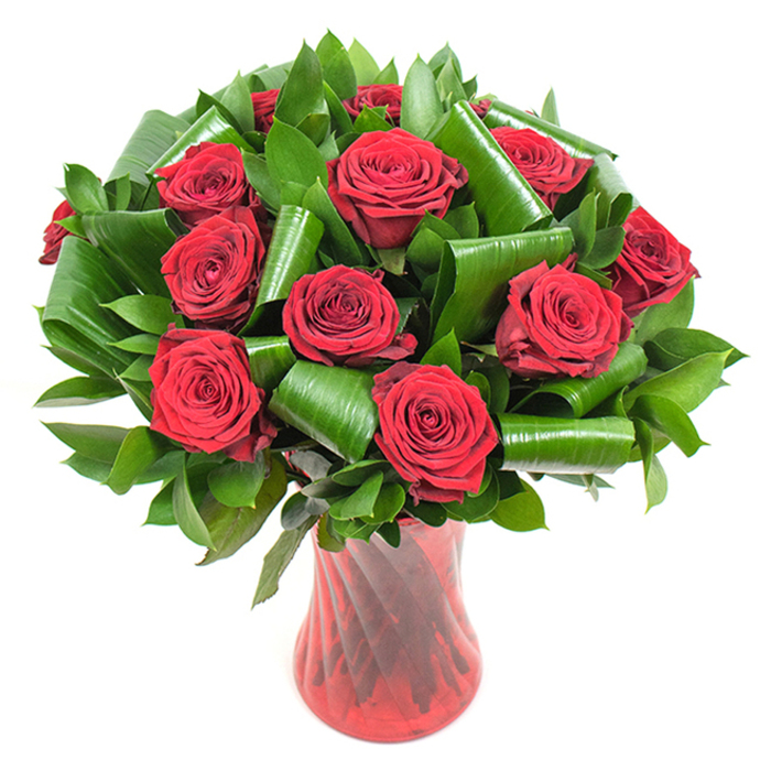 A sumptuous arrangement of a dozen or so deep crimson roses is presented here as a study in romantic texture and structure. Each rose is fully open, its velvety petals tightly spiralled at the centre then unfurling to soft, layered edges that catch the light and reveal subtle tonal shifts from bright scarlet to rich burgundy. Around and between the rose heads is abundant dark green foliage: smaller glossy leaves provide a plush, verdant backdrop while larger, brighter green leaves-styled in rolled, cylindrical loops that recall folded Aspidistra-create sculptural frames for individual blooms. These deliberate leaf shapes add modern architecture to a classic bouquet, offering movement and support that keeps the composition both airy and full. The flowers are anchored in a tall, translucent ruby-red glass vase whose faintly twisted ribs and slender profile throw vertical highlights under soft, even lighting, echoing the stems and enhancing the sense of height. Placed against a clean white field, the intense reds and greens become the sole focus, emphasising freshness and craftsmanship. As a Hook florist I'd mention that this arrangement, crafted at Flower Delivery Hook near the station and the High Street, carries the kind of presence suited to anniversaries, proposals, or any occasion calling for a clear declaration of love. Note the gentle perfume lifting from the blooms, the contrast between satin petals and glossy leaves, and the way the vase's rich ruby tone completes the composition with an elegant echo of the roses' depth.