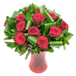 A lush, rounded bouquet of a dozen or more deep crimson roses fills the frame, each bloom a perfect cup of velvety petals spiralling inward toward a darker, wine-toned heart. The petals are shown in full blossom, their soft, satiny texture apparent even without touch, and a gentle, classic rose scent seems to rise from the tightly clustered heads. Deep, glossy green foliage surrounds and cushions the roses - smaller, lance-shaped leaves that read like Ruscus or Salal form a dense, verdant backdrop while larger, emerald leaves have been expertly folded and rolled into cylindrical, architectural loops that cradle and lift the blooms. Those sculpted leaves give the arrangement a contemporary, almost sculptural rhythm that balances the old-world romance of the rose. The bouquet sits in a tall, translucent ruby red glass vase whose subtle ribbed twist catches and reflects the soft, even studio light, creating vertical highlights that echo the stems within. The vase's narrower waist and gently flared lip lend an elegant silhouette, anchoring the composition against a clean white background so the saturated reds and greens read with strong clarity. The overall feeling is one of refined passion and quiet luxury - a centrepiece suited to an anniversary, Valentine's gesture, or a thoughtful celebration delivered locally around Hook village and the High Street. As a florist describing this to a customer, I'd note the freshness of the blooms, the contrasting textures, and the way the lighting reveals every velvety fold - an arrangement that reads as timeless romance with a modern twist.