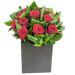 In this elegantly modern composition, deep crimson roses form the arresting centre-each bloom a velvety cup with petals unfurling in soft, generous spirals that catch the light like satin. The roses are cradled by broad, glossy green leaves that have been carefully looped and curled, their surfaces catching bright highlights and creating a rhythmic pattern of movement and shadow around the blooms. The arrangement sits in a crisp, matte black cube that gives the bouquet a grounded, architectural quality; the square lines of the vessel contrast the organic curves of petals and foliage, emphasising the composition's contemporary poise. Lighting in the photograph is clear and even, lifting the crimson tones until they glow with a tactile, almost touchable richness while the greens remain fresh and luminous. If you lean in, there's the imagined scent of classic rose-warm and full-balanced by the cool green notes of freshly cut leaves, a subtle fragrance that feels both comforting and refined. As a florist who often arranges for celebrations on Hook High Street and deliveries to homes near the station, I see this bouquet as an intentional statement piece: suited to an anniversary table, a romantic milestone, or an urban living room in need of a bold focal point. The craftsmanship is quiet but confident-the leaves sculpted to frame each rose without overpowering it-so that the overall effect is of passionate restraint, an elegant declaration of feeling presented with modern clarity and local knowingness about what lifts a room in Hook and the surrounding villages.